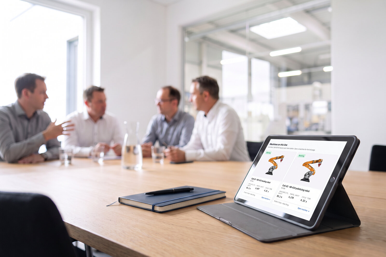 A tablet on a meeting-room table showing the machines on a line - two robot cells, each with its availability, parts and cycle time for the shift - managers out of focus behind. Illustrative photo, demo data.