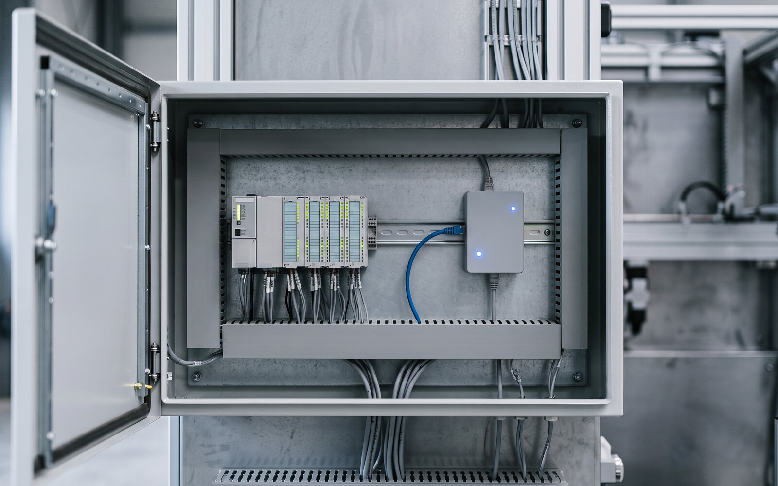 Inside the open control cabinet of a machine: a compact industrial gateway with its blue status LED, wired with a blue Ethernet cable to the modular PLC on the DIN rail. Illustrative photo.