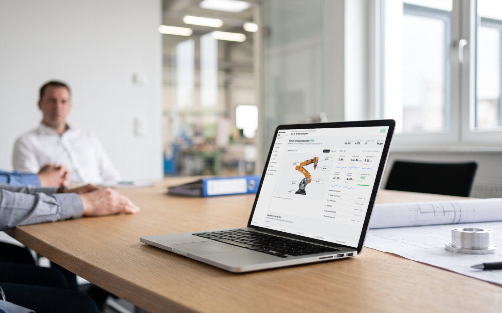 A laptop on a meeting-room table showing a machine page under the customer's own brand, live values pinned on the 3D model of a robot, a technical drawing beside it. Demo data, illustrative photo.