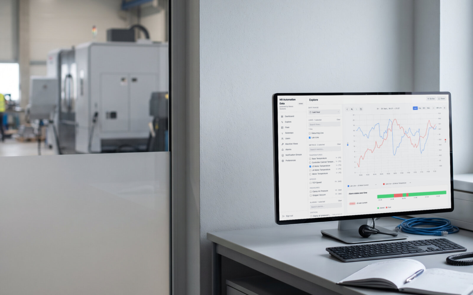 A service engineer's desk by the assembly hall, the monitor showing an Explore chart under the customer's own brand: two robot signals on their own axes, an alarm timeline below. Demo data, illustrative photo.
